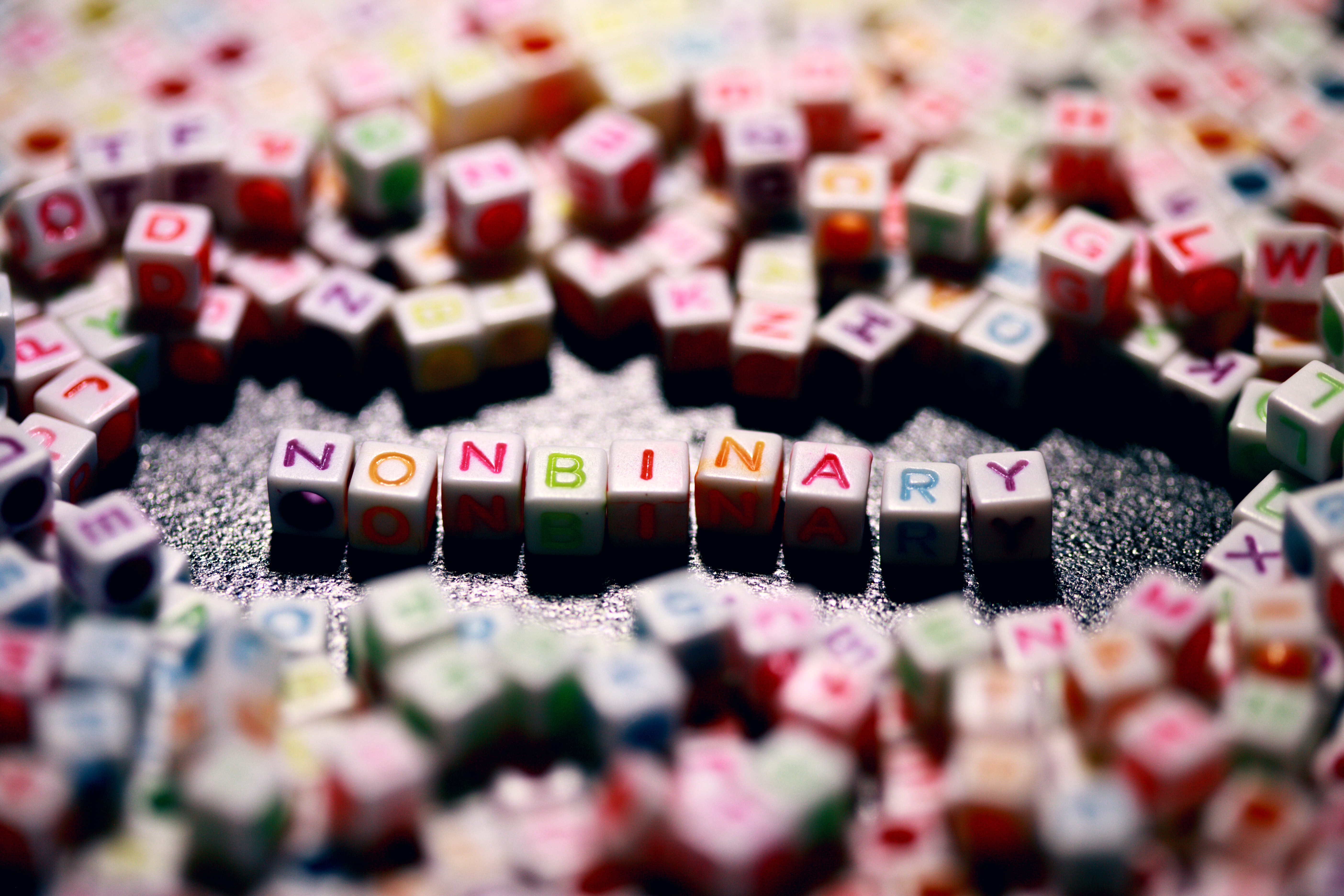 multicolored beaded letters on a surface, the center of the photo has the word nonbinary spelled with different colors.